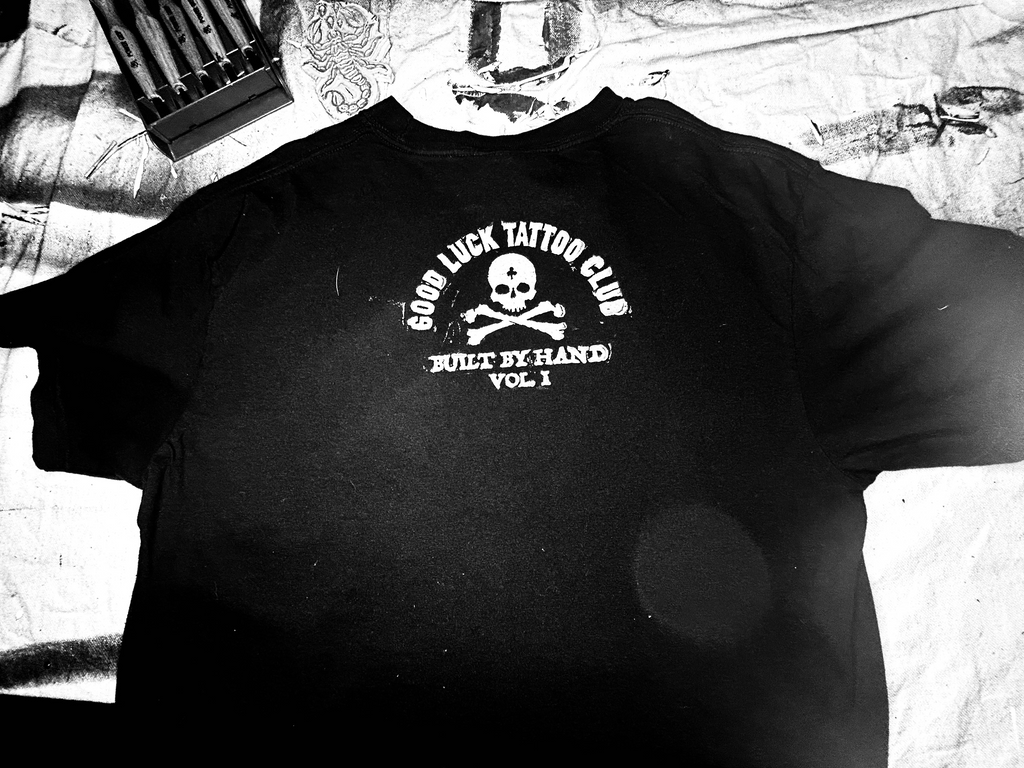 Black hoodie with a logo on the chest, worn by a person, against a tattooed arm background.